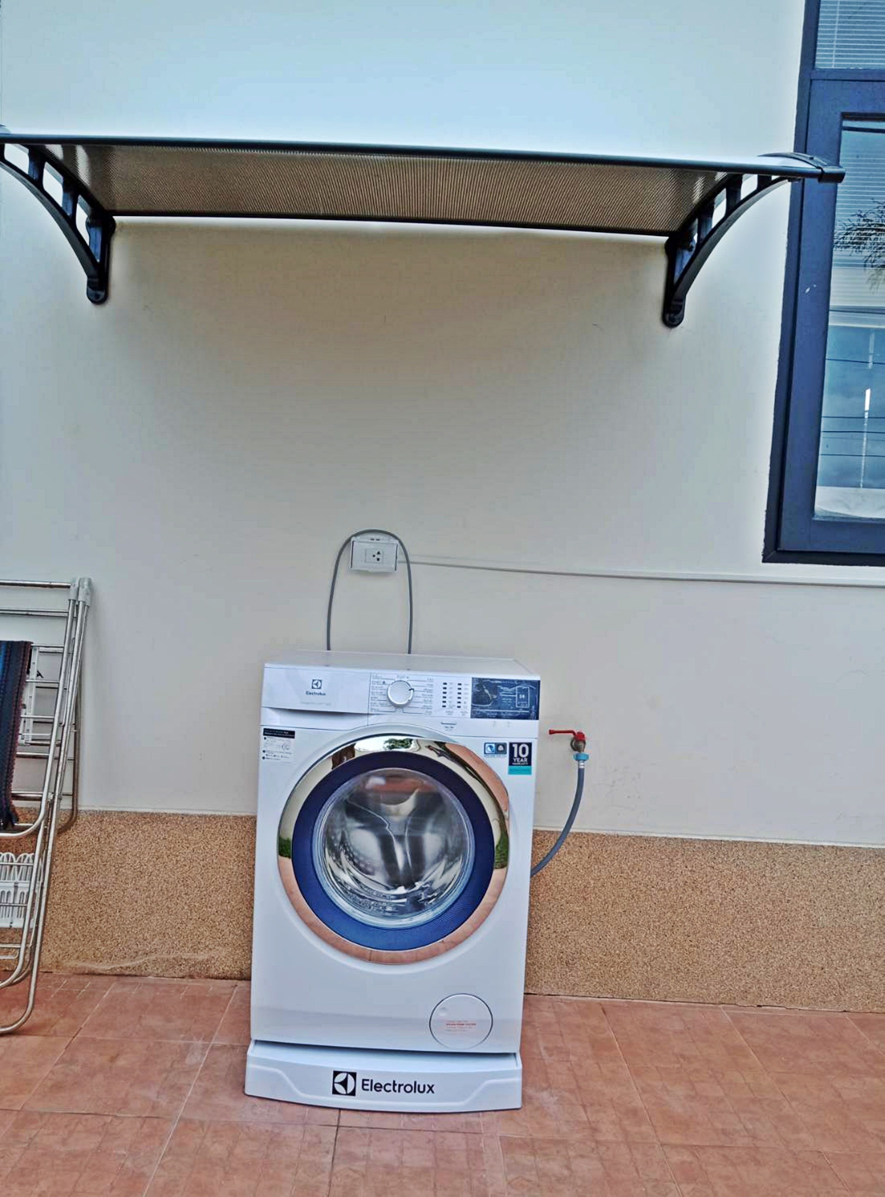 A front-loading washing machine with a control panel, connected to a power outlet, and a metal drying rack to its left.