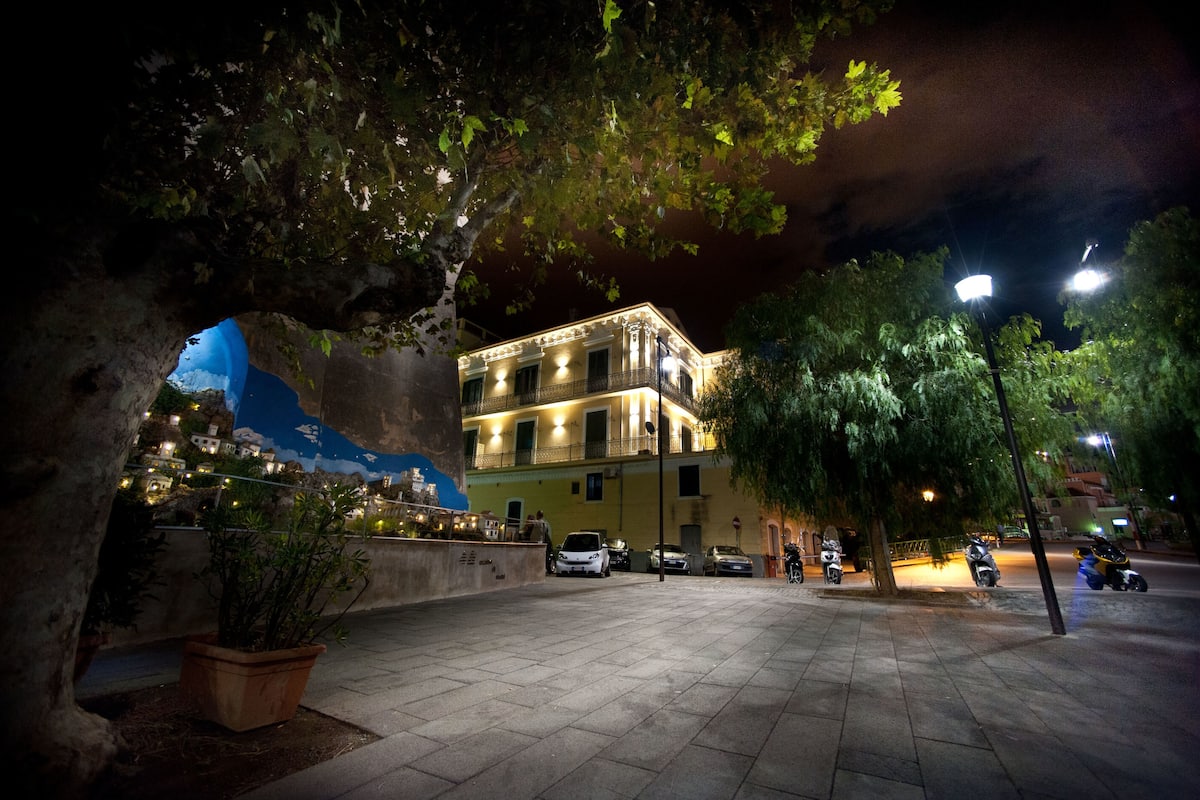 Escena urbana nocturna con un edificio que tiene balcones y una calle bordeada de árboles.