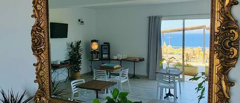 A room with a large ornate mirror, a dining area with white chairs, a small table, and a view of the sea through large windows.