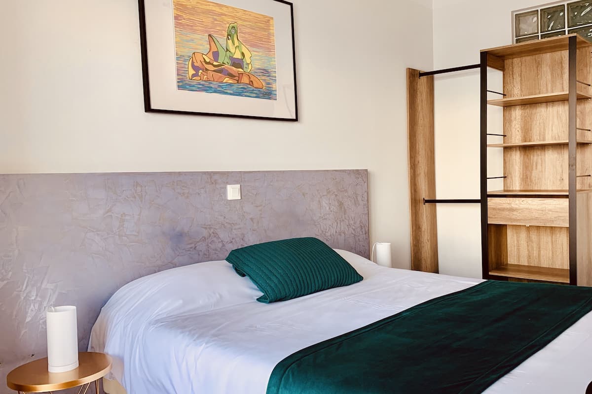 A bedroom with a bed, a bedside table, a wall-mounted shelf, and a framed artwork.