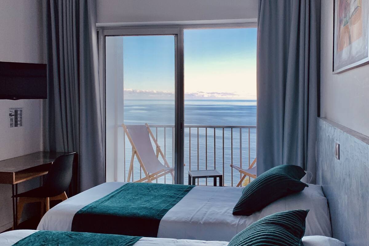 A hotel room with a bed, a desk, and a chair. There is a balcony with a view of the sea.