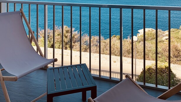 Un balcón con dos sillones de descanso y una mesita pequeña, con vistas a la costa, el mar y terreno rocoso.