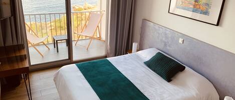 A hotel room with a bed, a view of the sea, a balcony with chairs, and a painting on the wall.
