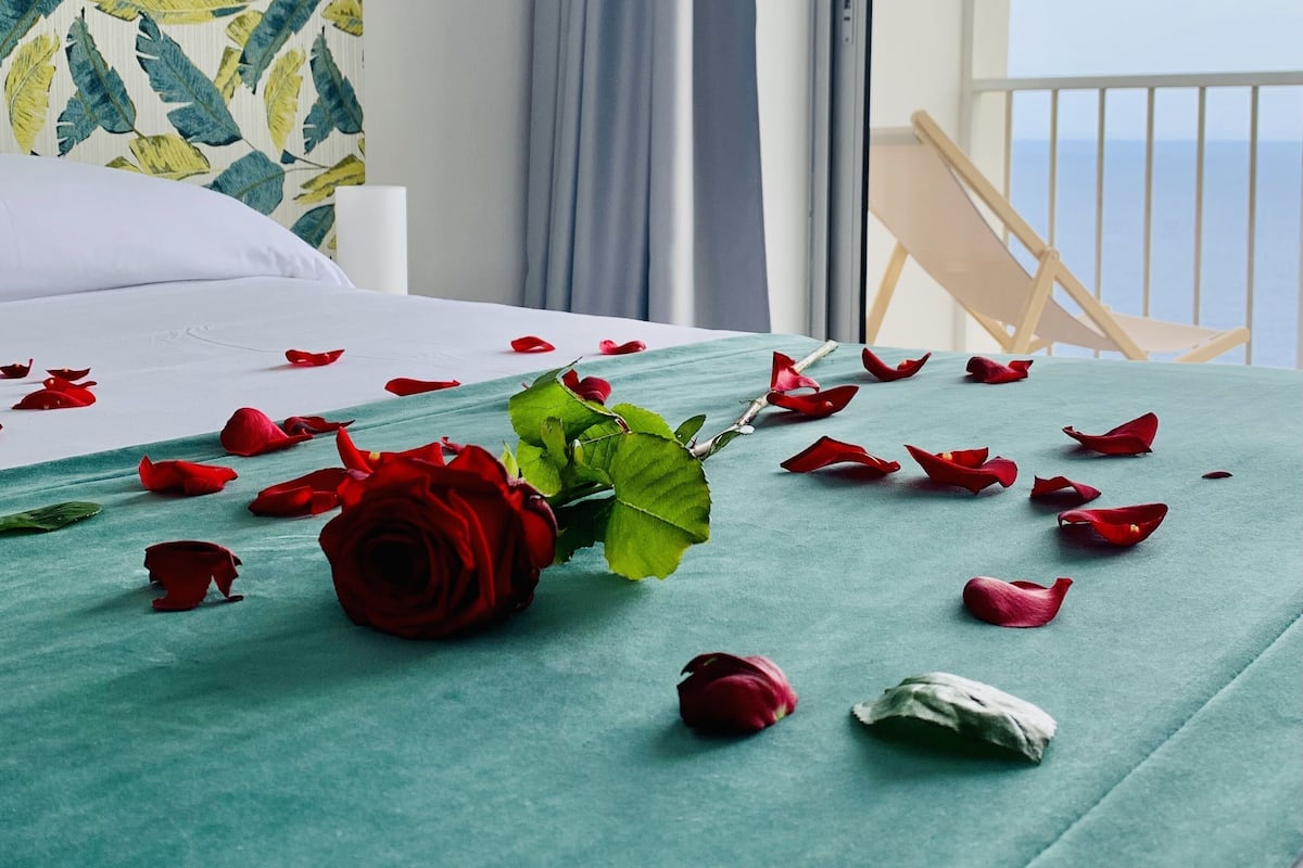 A neatly made bed with a green bedspread, a single red rose, and scattered rose petals.
