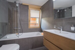 A modern bathroom with a glass-enclosed shower, a large bathtub, a double sink vanity, and a window with blinds. - Ferienhaus Felicé (Zermatt)