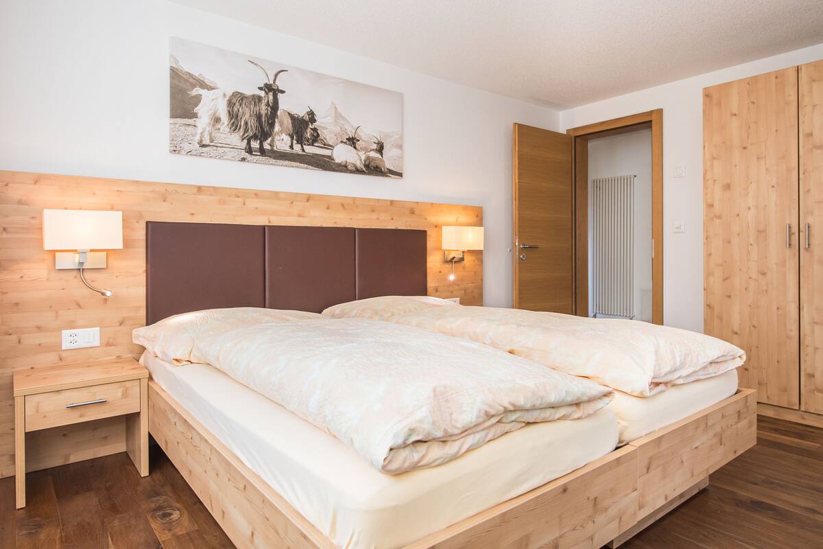 A double bed with a wooden headboard, two bedside tables with lamps, a wall-mounted picture, and a wooden wardrobe.