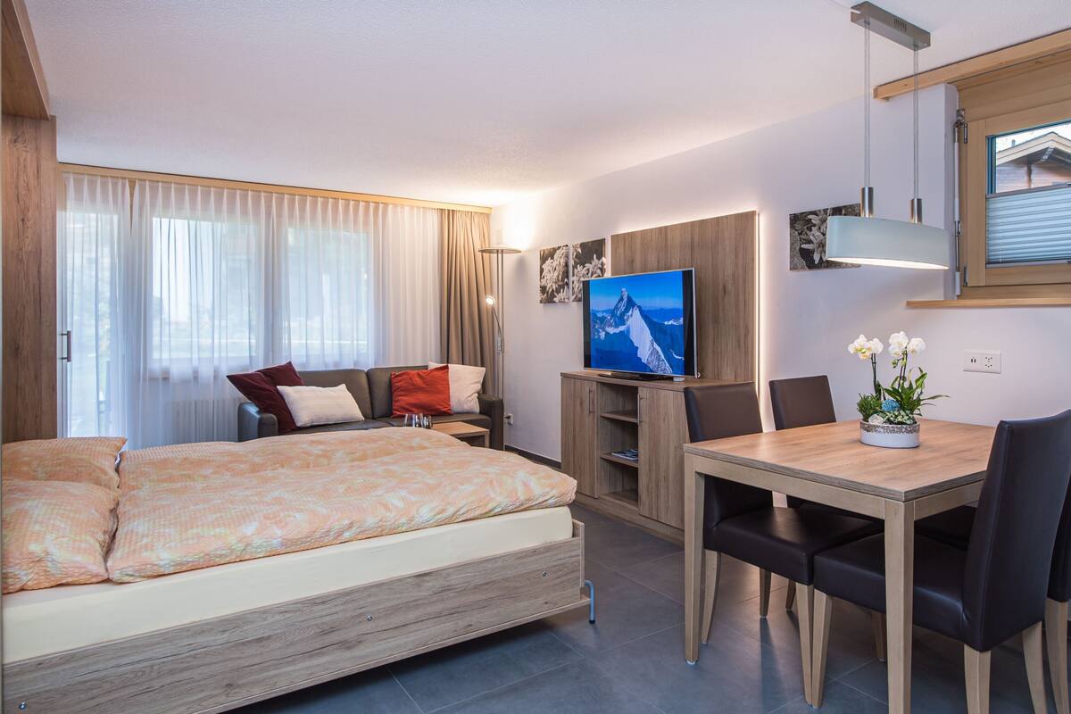 A modern hotel room with a bed, a TV, a dining table, and a small plant.