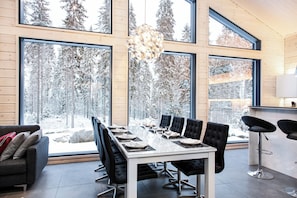 Uma sala de jantar moderna com uma mesa comprida, cadeiras estofadas pretas e grandes janelas com vista para uma floresta coberta de neve.