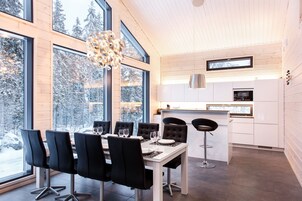 Un comedor moderno con una gran mesa y sillas, una cocina con armarios blancos y ventanas amplias que ofrecen vistas a árboles cubiertos de nieve.