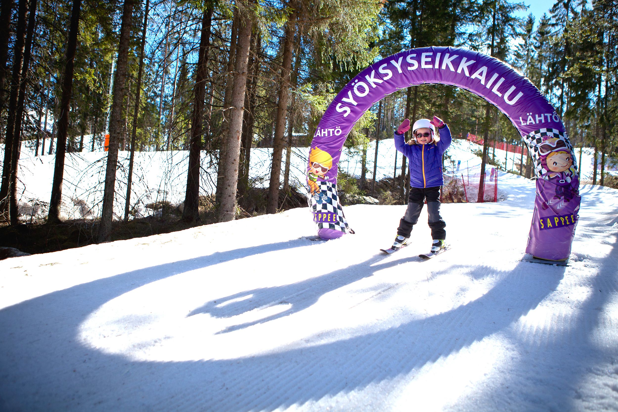 A snowy ski area with a purple archway labeled "SYÖKSYSEIKKAILU" and a person in a blue jacket standing under it.