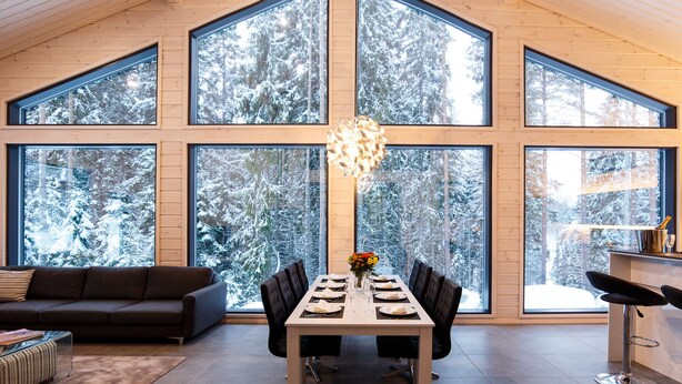 A modern dining area with a large table and chairs, large windows offering a view of snow-covered trees. - Villa Borealis (Pälkäne)