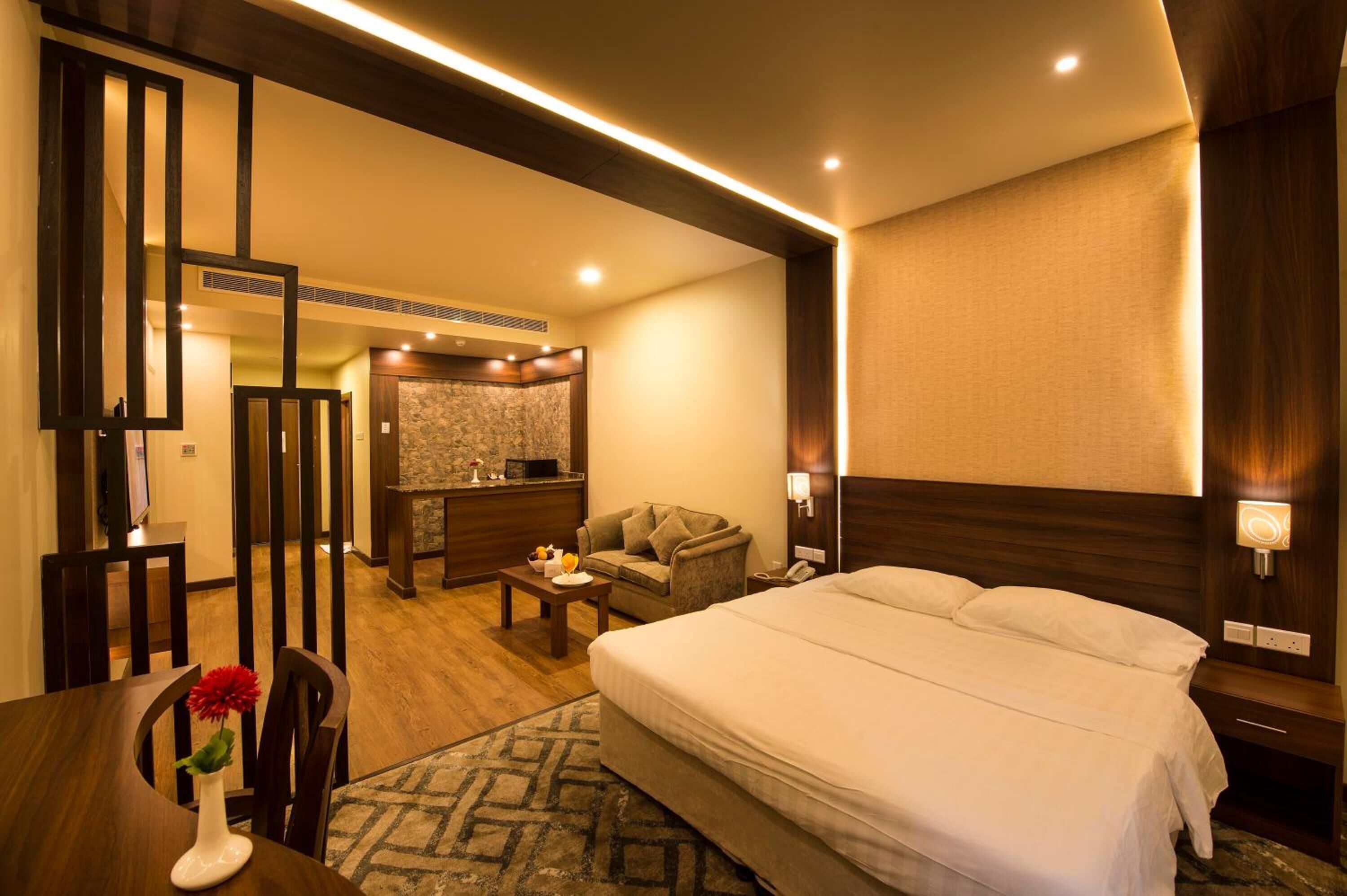 A hotel room with a large bed, a desk, a chair, a sofa, and a small kitchen area.