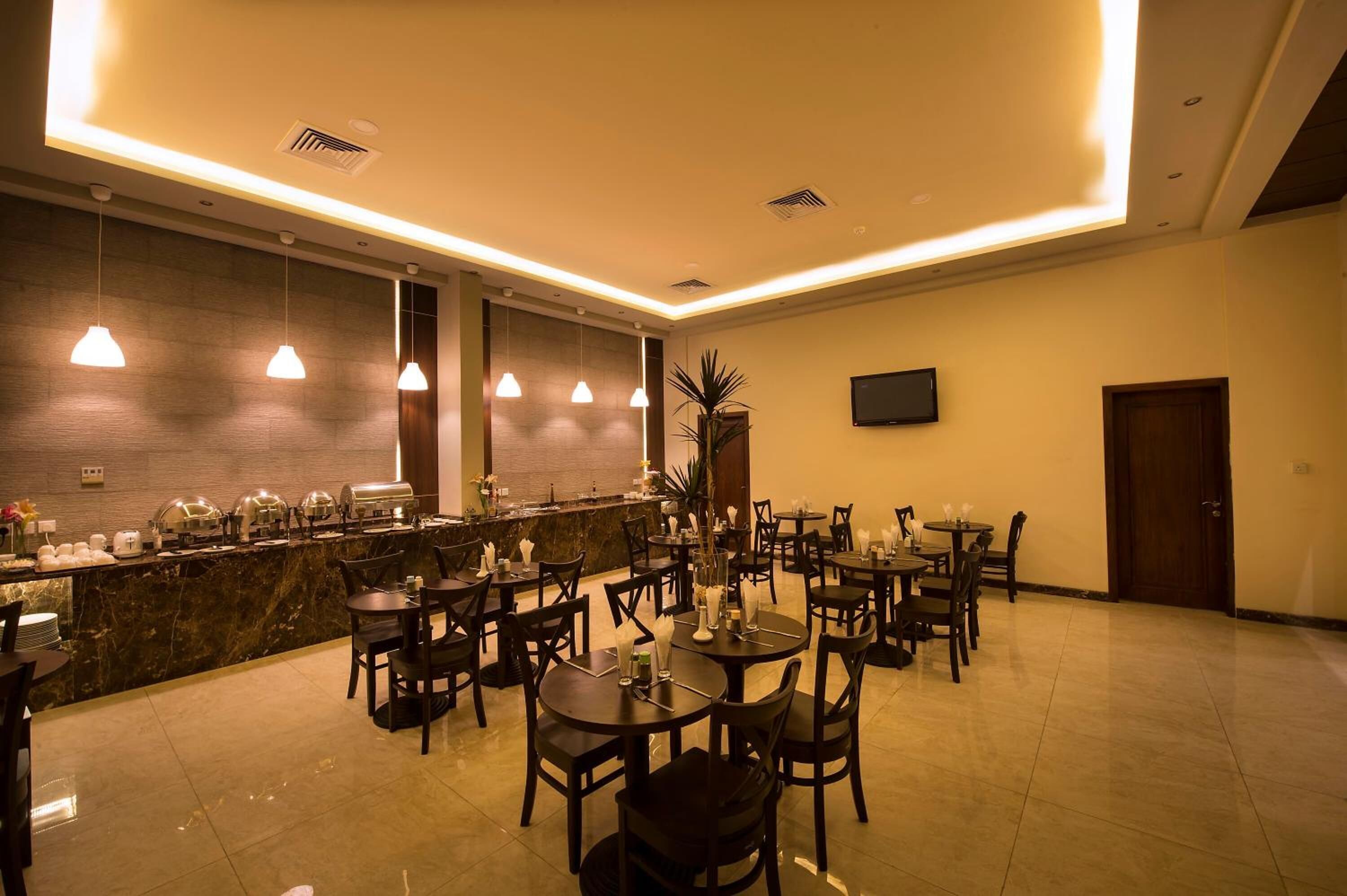Uma sala de jantar de hotel com mesas e cadeiras redondas, um balcão de buffet e uma televisão fixada na parede.