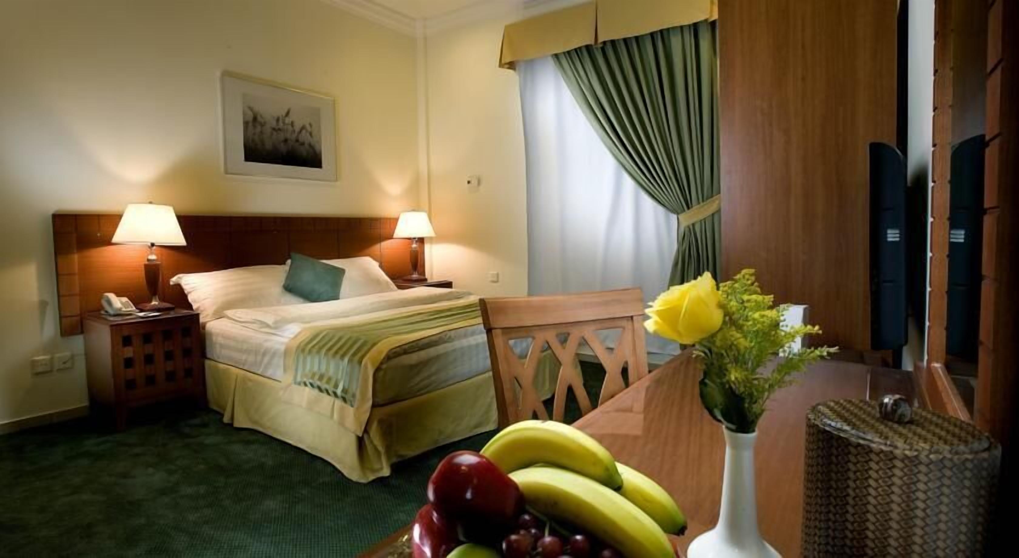 A hotel room with two beds, a desk with fruits, a vase with flowers, a painting on the wall, and a window with curtains.