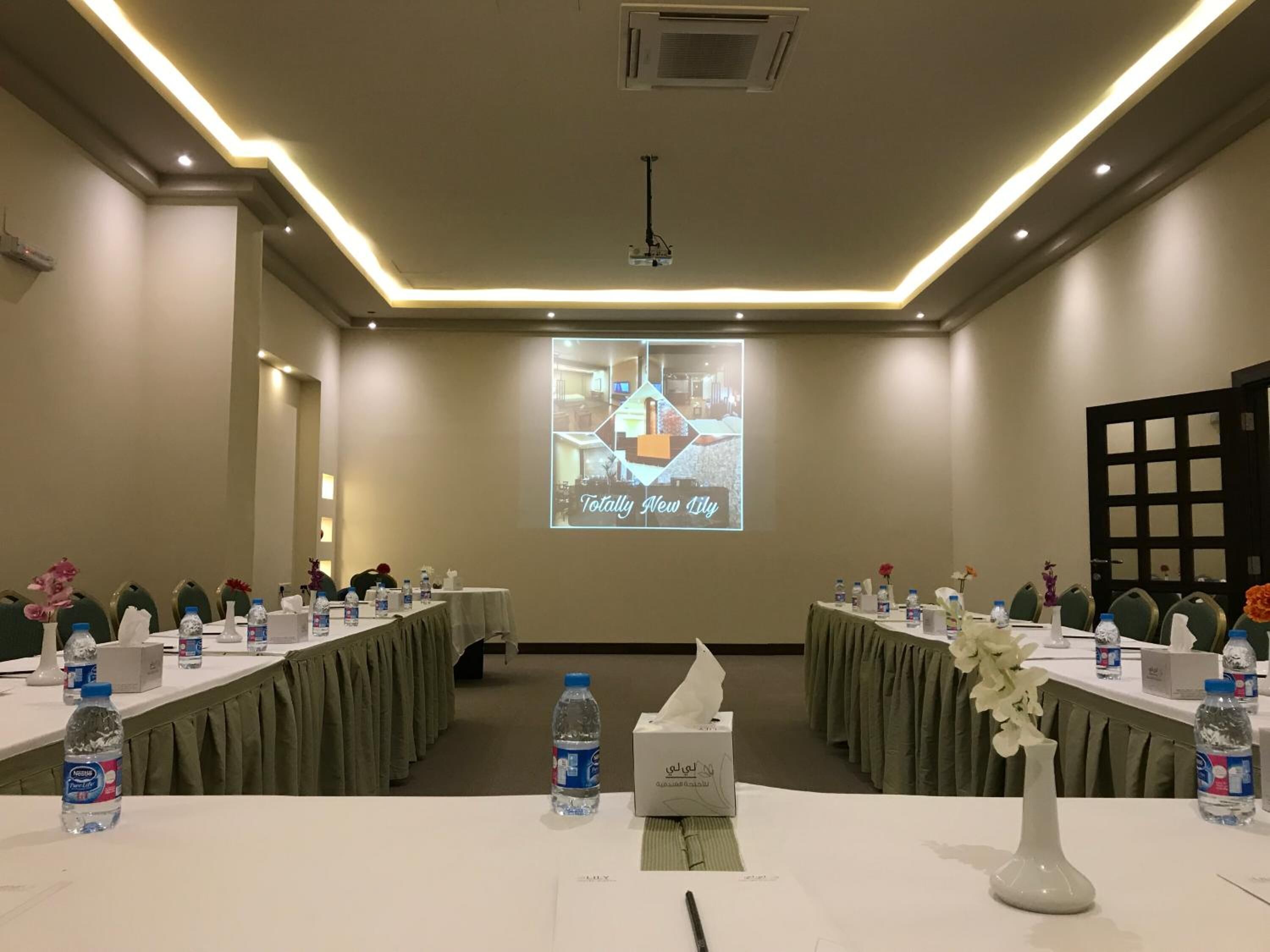 Uma sala de conferência com mesas preparadas para uma reunião, equipada com garrafas de água, lenços de papel e uma tela de projeção exibindo uma apresentação.