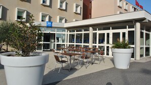 Een terras met tafels en stoelen voor een gebouw met veel ramen en een bord waarop 