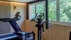 Een sportschool met een loopband en een elliptische machine, grote ramen met uitzicht op bomen, en een wandlamp.