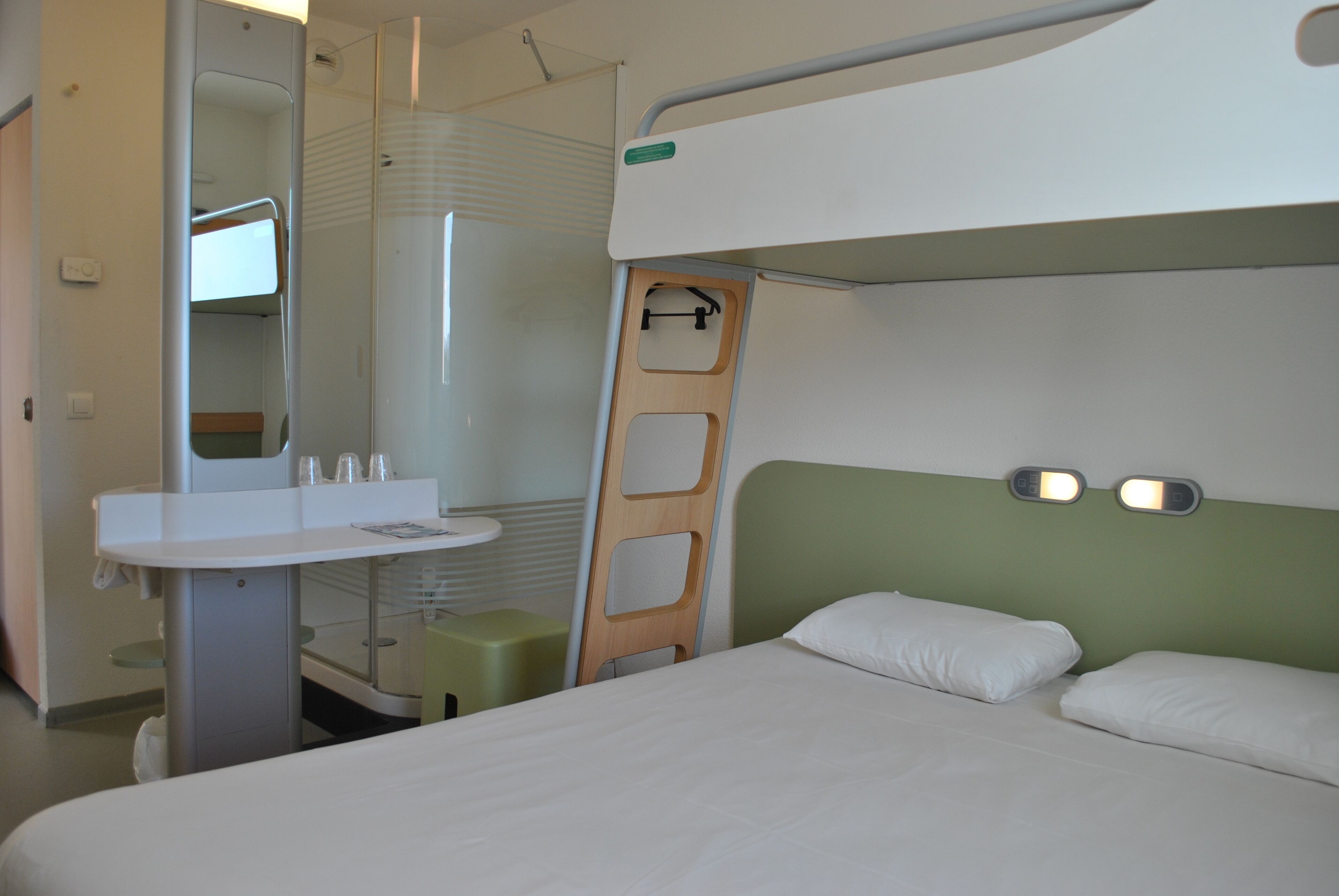 Une chambre d’hôtel compacte comprenant un lit superposé, une petite table de chevet, un miroir et un espace douche.