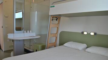 Une chambre d’hôtel compacte comprenant un lit superposé, une petite table de chevet, un miroir et un espace douche.