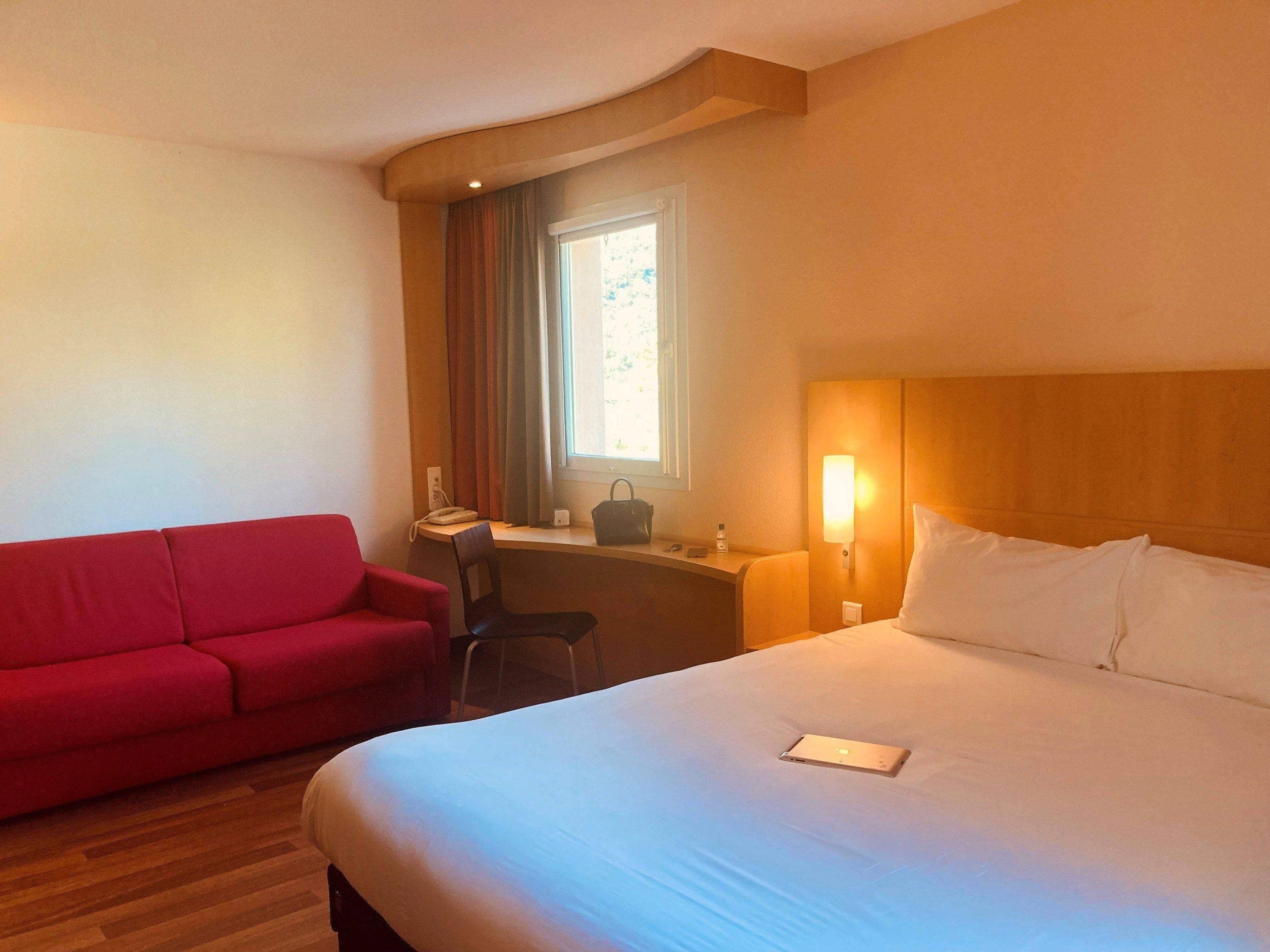 A hotel room with a bed, a red sofa, a desk with a chair, a window with curtains, and a wall-mounted lamp.