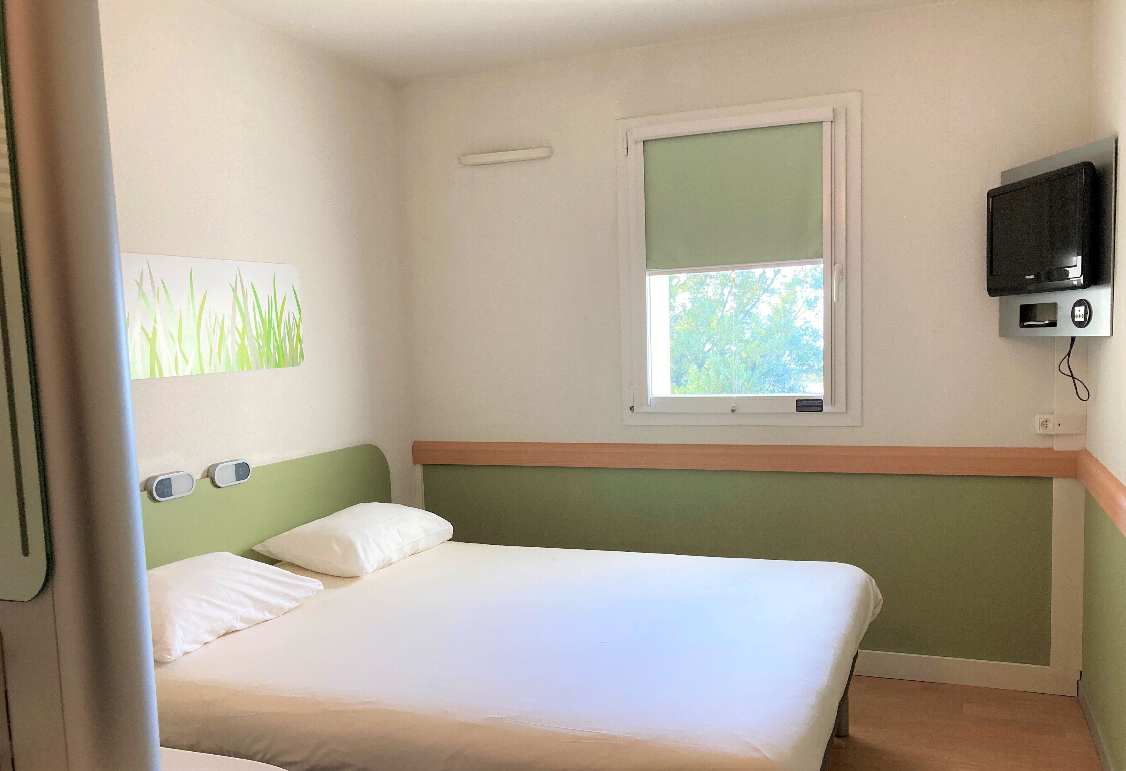 Une chambre d’hôtel avec un lit, une fenêtre, un téléviseur et un interrupteur fixé au mur.