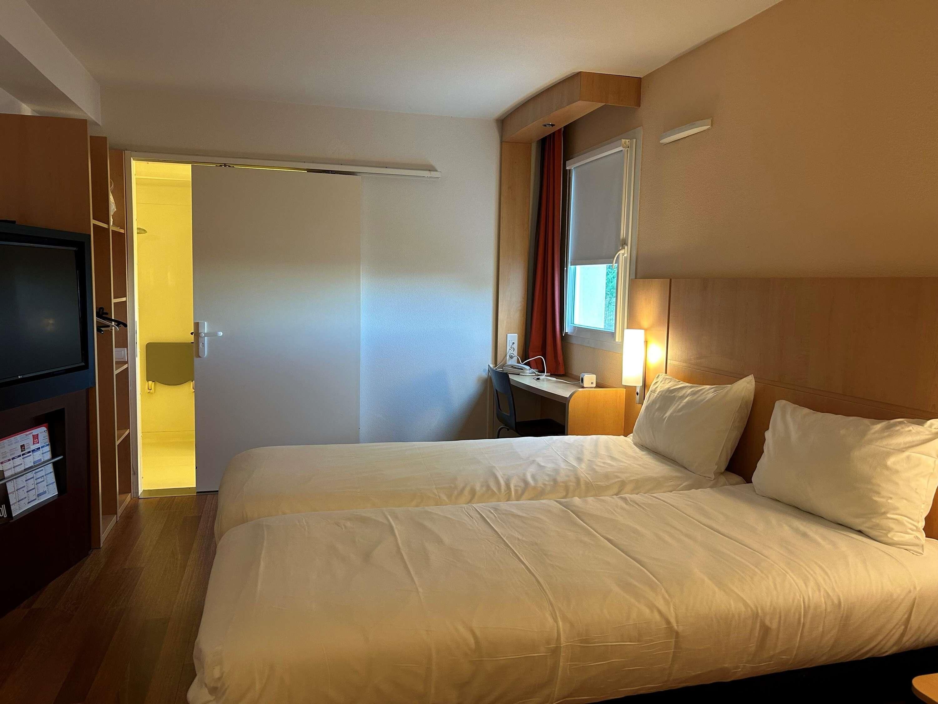 A hotel room with two beds, a desk with a chair, a television, and a window with curtains.
