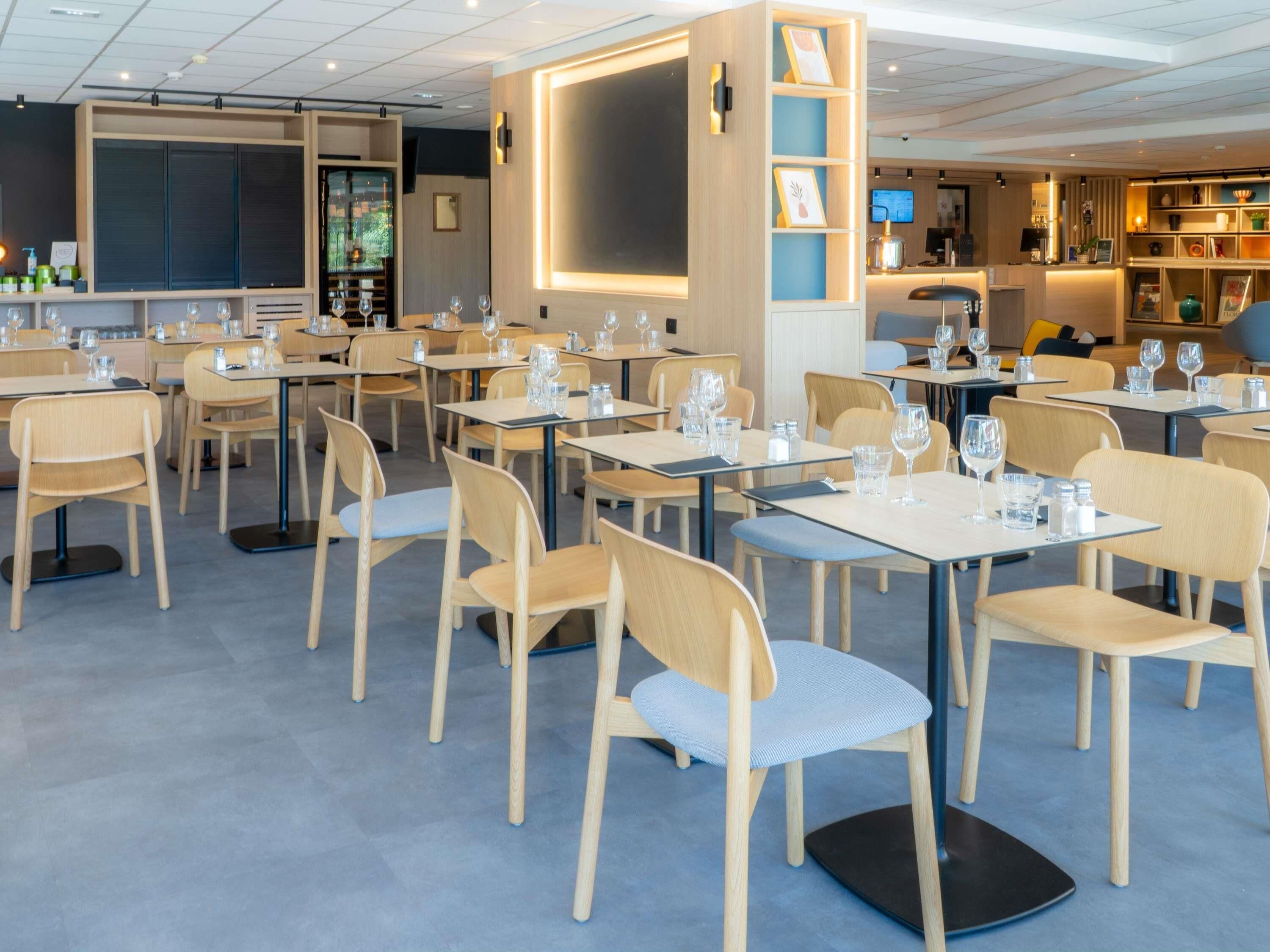 A modern restaurant with multiple tables and chairs, a bar area, and a counter with a digital screen.