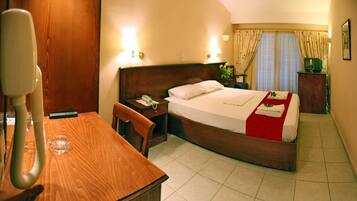 A hotel room with a wooden bed, a desk, and a chair. There is a telephone on the desk and a television on a wooden cabinet.