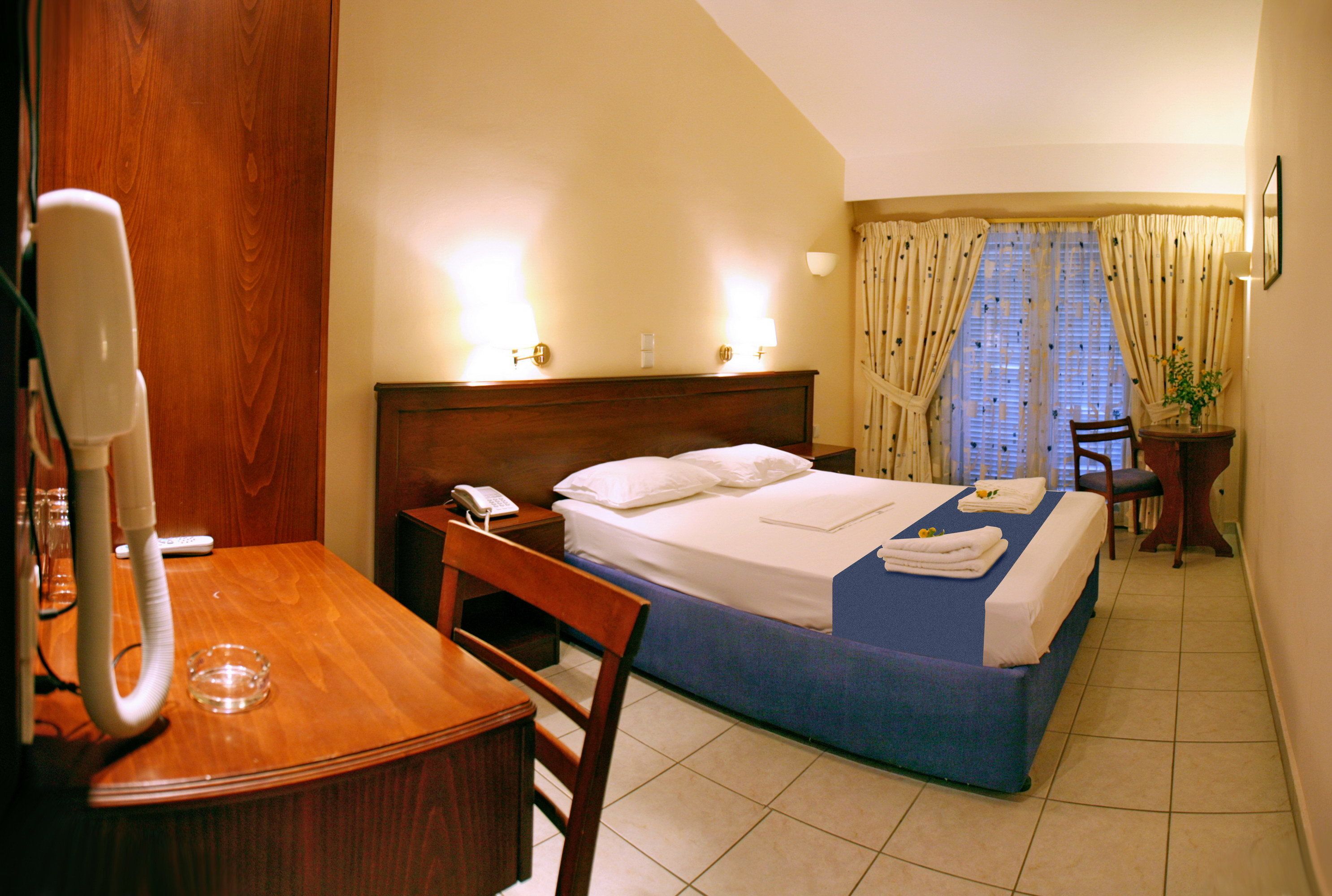 A hotel room with a wooden desk, a chair, a bed with a blue bedspread, a bedside table with a lamp, and a window with curtains.