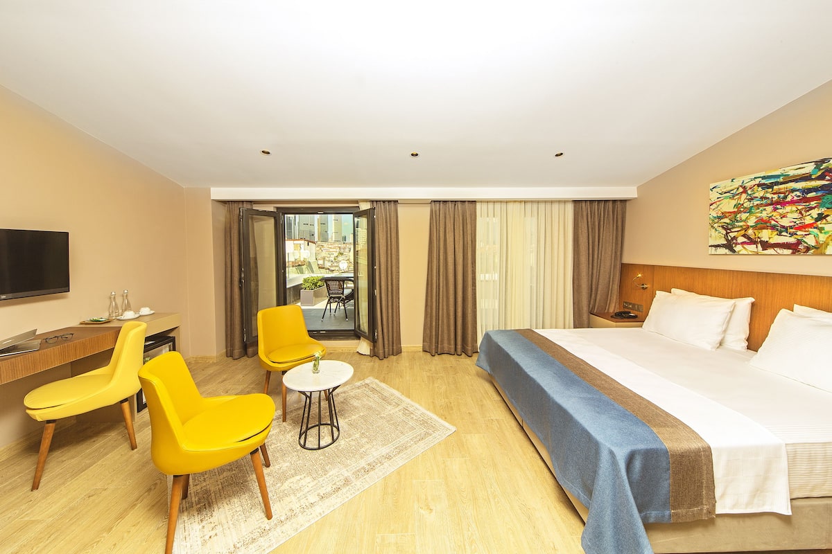 Quarto de hotel com uma cama, uma escrivaninha, duas cadeiras amarelas, uma mesinha e vista para o exterior.