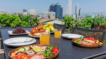Une table à manger installée sur un toit, offrant une vue panoramique sur la ville, avec des assiettes de nourriture, des fruits et un verre de jus.