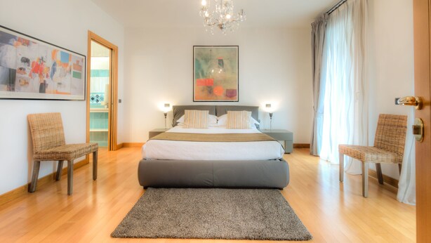 A modern bedroom with a large bed, two bedside tables, a chandelier, and a view of a cityscape through a window. - Domus (Ravenna)