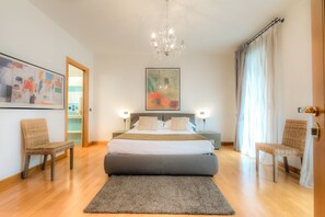 Un dormitorio moderno con una cama grande, dos mesitas de noche, una araña y vistas a un paisaje urbano a través de una ventana.