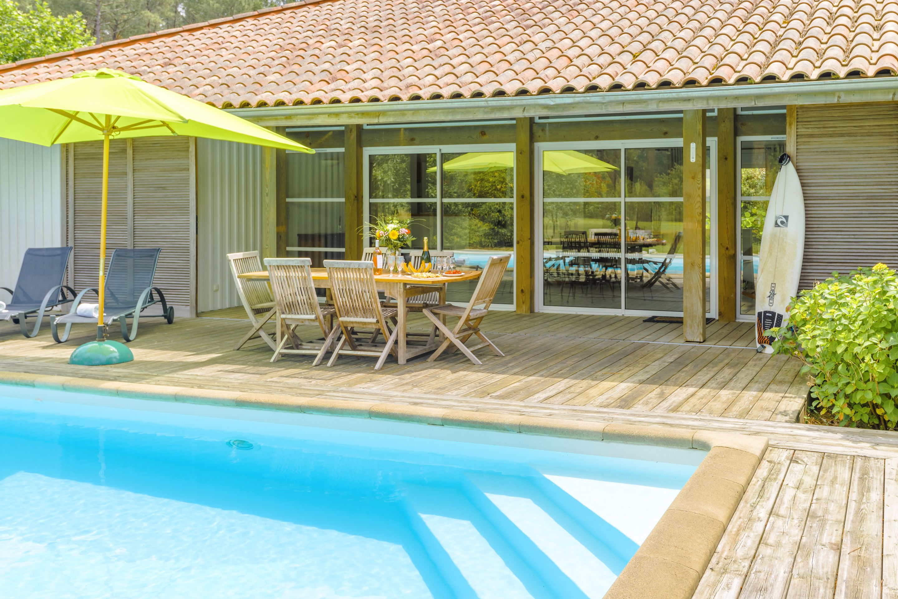 Zona de piscina con terraza de madera, una mesa con sillas y un paraguas amarillo.