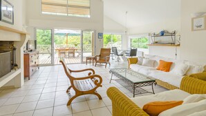 Ein großzügiger Wohnbereich mit einem großen Glasschaufenster, das auf eine Terrasse im Freien führt, eine Couch, ein Couchtisch und Holzstühle.