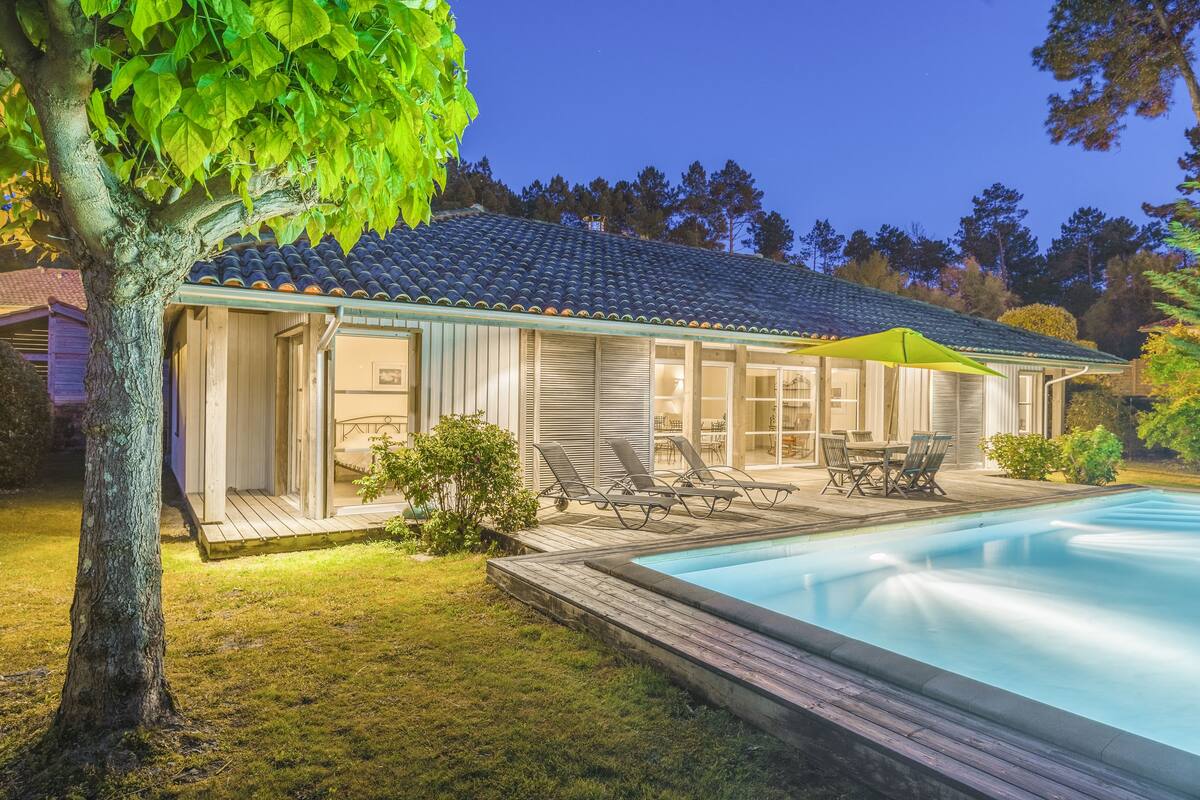 Ein modernes Haus mit Swimmingpool, einer Terrasse mit Gartenmöbeln und einem großen Baum im Vordergrund.