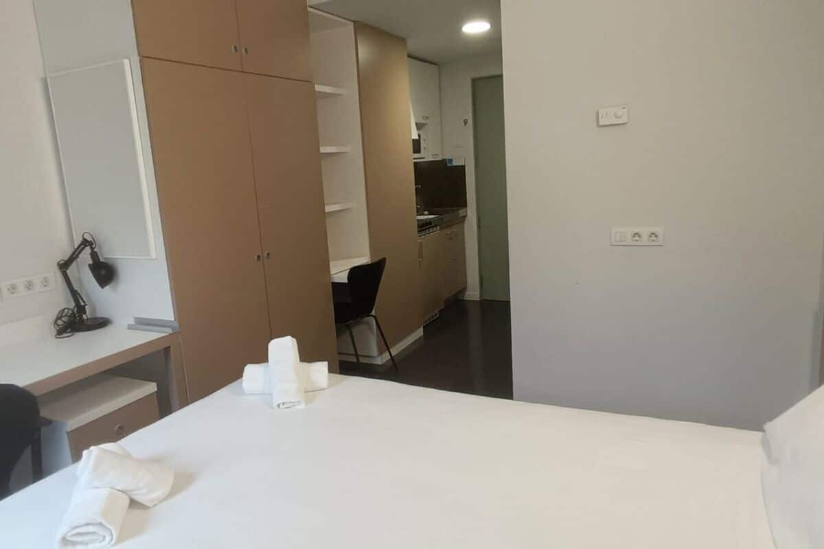 Hotelkamer met een bed, een bureau met lamp, een stoel en een kitchenette op de achtergrond.