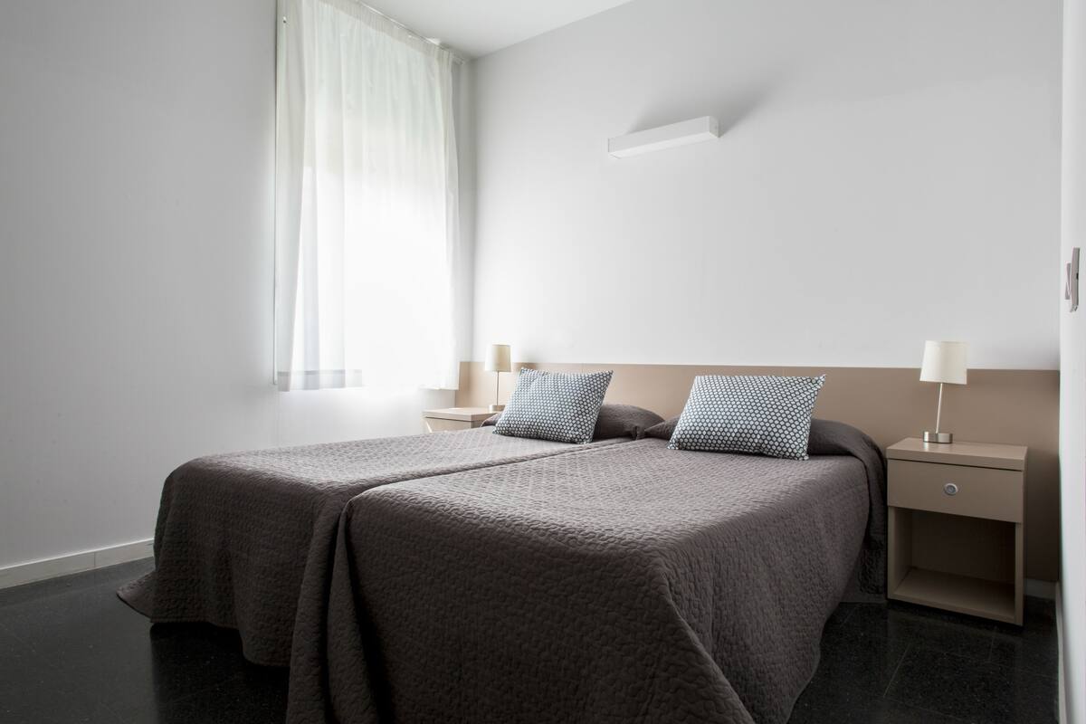 Een moderne hotelkamer met een tweepersoonsbed, twee nachtkastjes en een raam met dunne gordijnen.