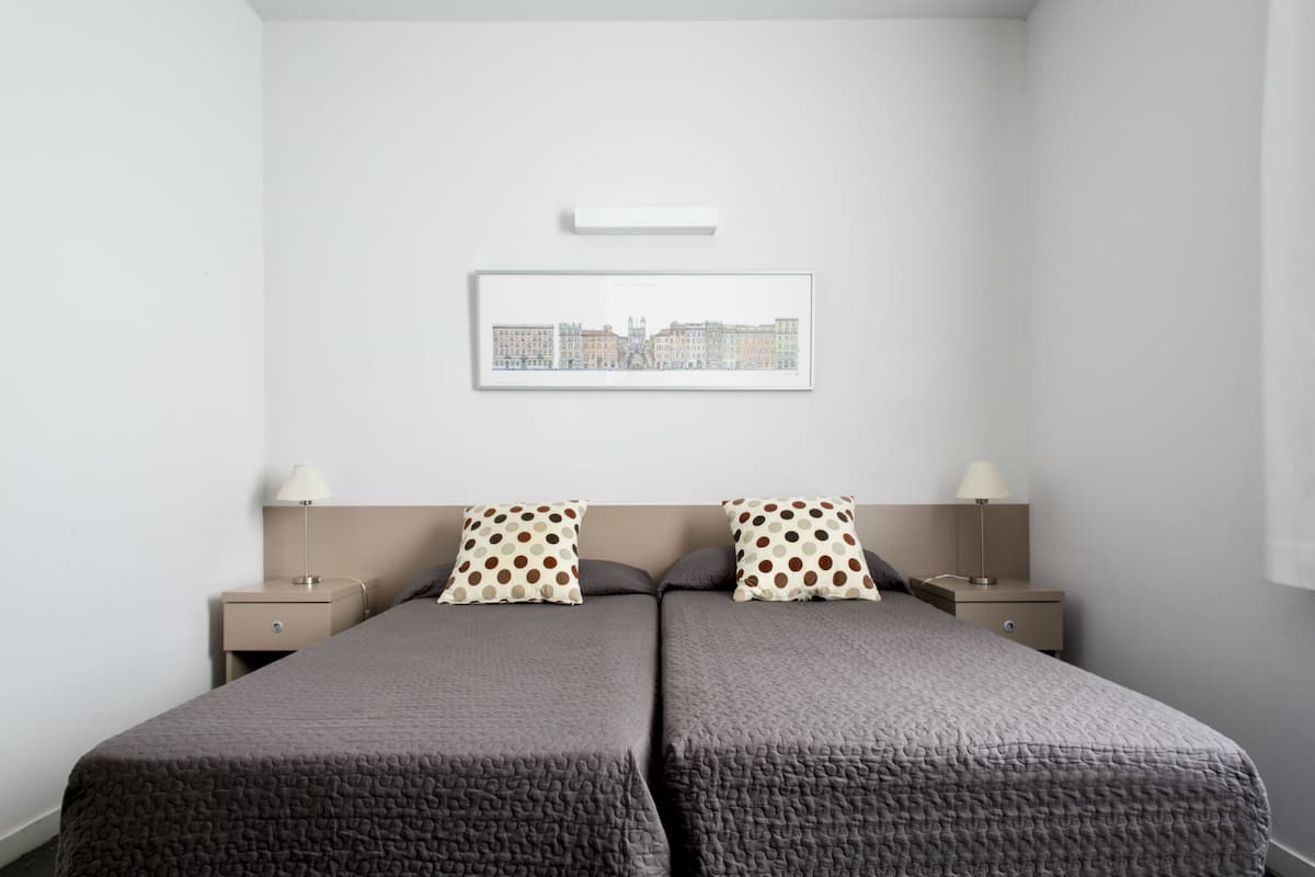 Een tweepersoonsbed met grijze beddengoed, twee nachtkastjes met lampen en een lijst met een stadsgezicht aan de muur.