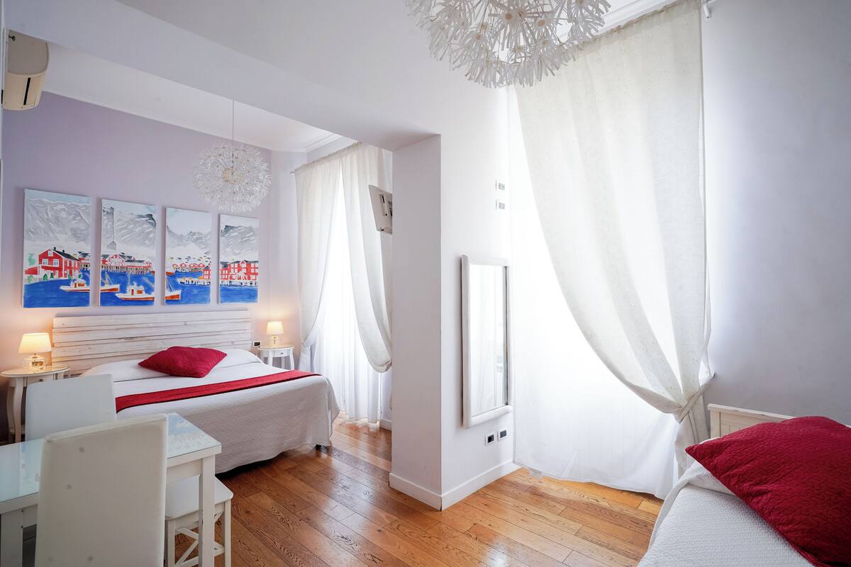 A hotel room with a bed, a desk, and a chair. There are paintings on the wall and a window with curtains.