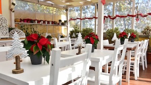 Un área de comedor al aire libre cubierta, con mesas y sillas blancas, decorada con flores de Pascua y pequeños árboles de Navidad.
