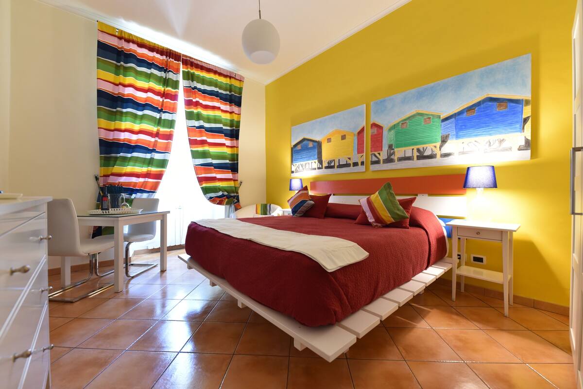 A bedroom with a bed, a desk, and a chair. There is a painting on the wall and a window with curtains.