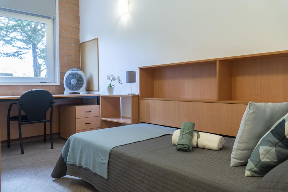Une chambre d’hôtel équipée d’un lit, d’un bureau, d’une chaise, d’un ventilateur et d’une fenêtre donnant sur les arbres.