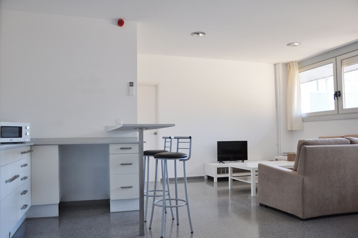 Une pièce moderne et bien éclairée, dotée d’une kitchenette, d’un coin repas avec une table blanche et deux tabourets de bar, d’un téléviseur à écran plat et d’un canapé beige.