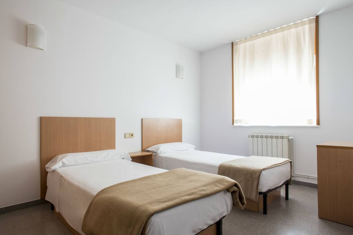 Une chambre d’hôtel avec deux lits simples, une tête de lit en bois, un radiateur et une fenêtre avec des rideaux.