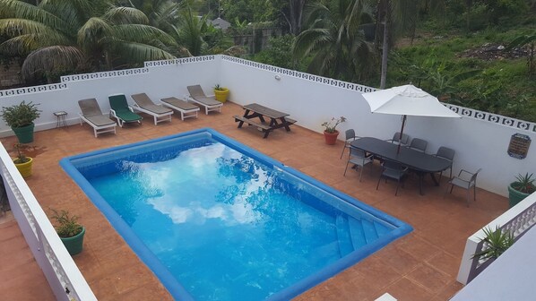 A rectangular swimming pool surrounded by lounge chairs and a table with chairs, set against a white wall with a decorative border.