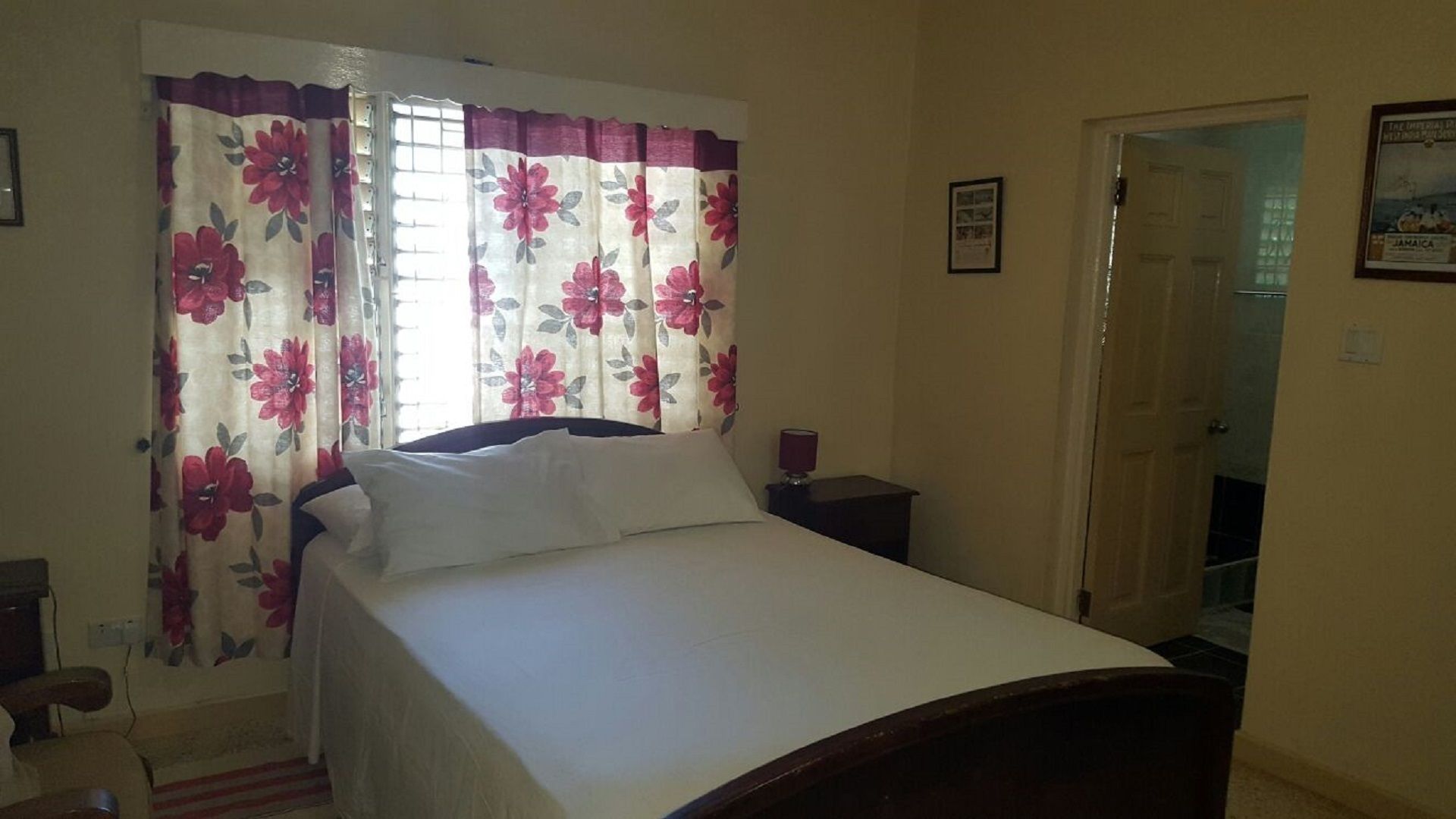 Une chambre à coucher avec un lit, des rideaux à motifs floraux, une table de chevet avec une lampe et une porte donnant sur une autre pièce.