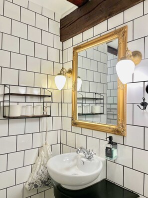 A bathroom with white tiled walls, a gold-framed mirror, a white sink, and a shelf with toiletries. - Hotel Hamngatan 27 (Falkenberg)