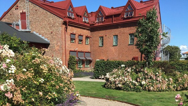 A brick building with a red roof, a gravel pathway, and flower beds with pink and purple flowers. - Hotel Hamngatan 27 (Falkenberg)