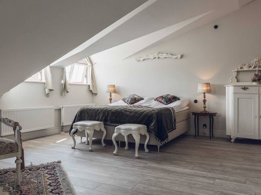 A bedroom with a sloped ceiling, a bed with a dark quilt, two bedside tables with lamps, a white wardrobe, and a patterned rug.